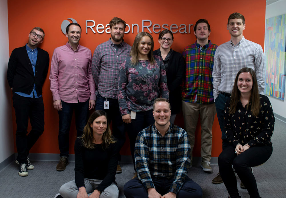 Two rows of individuals in front of orange Reason Research wall 