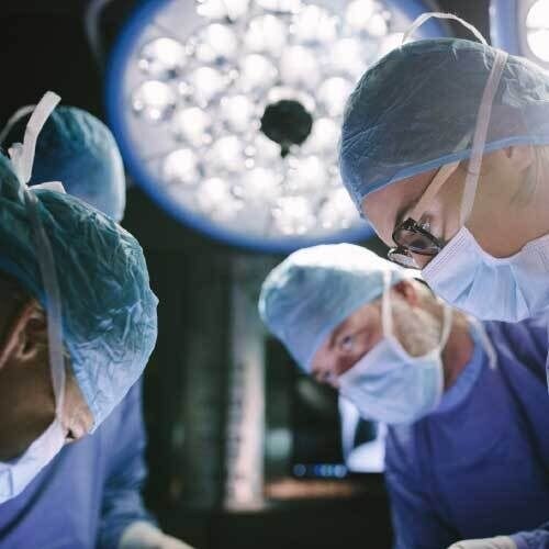 Doctors gowned up closely inspecting something in the OR with light behind them
