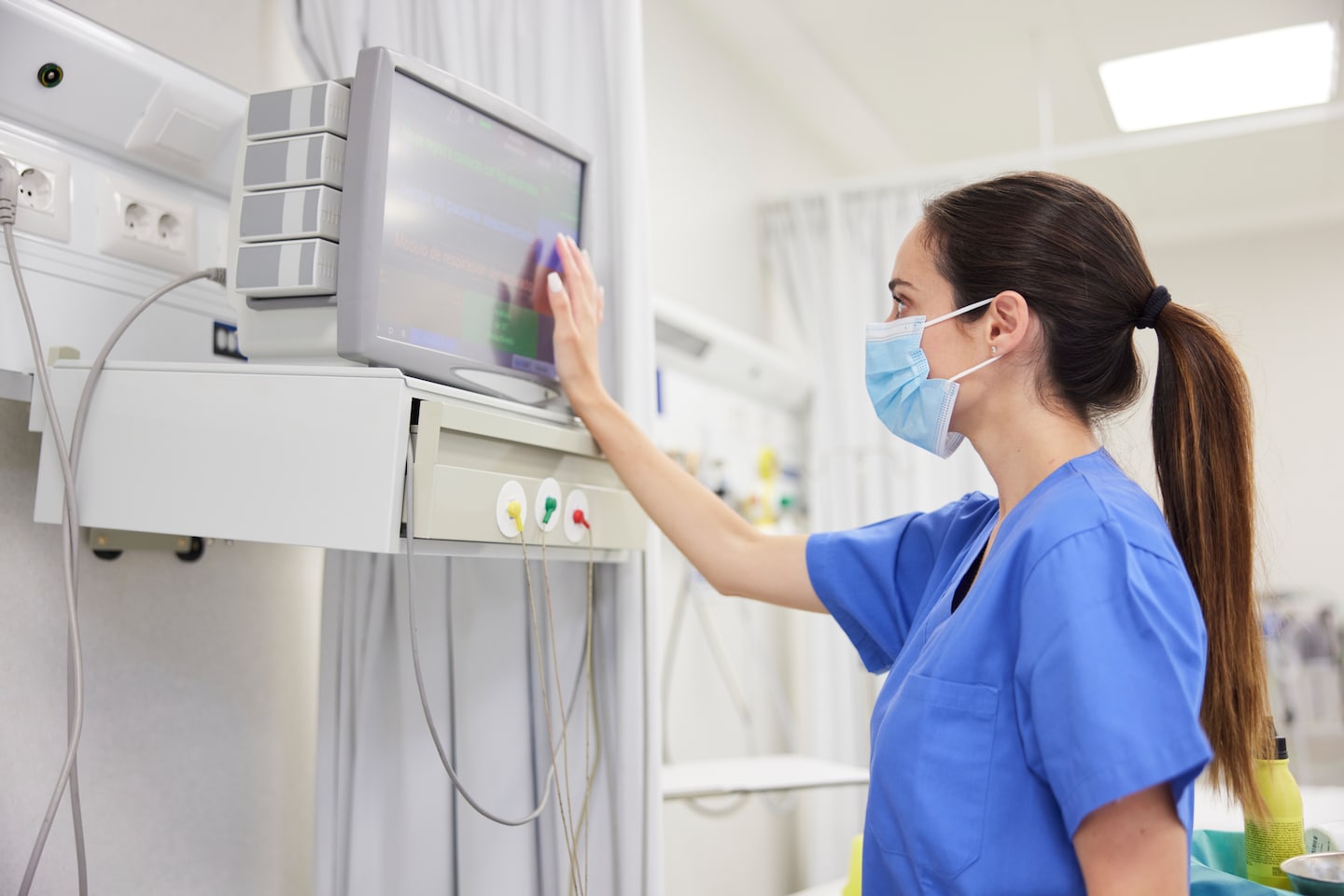 Young female healthcare professional with mask is touching patient monitoring screen in patient room