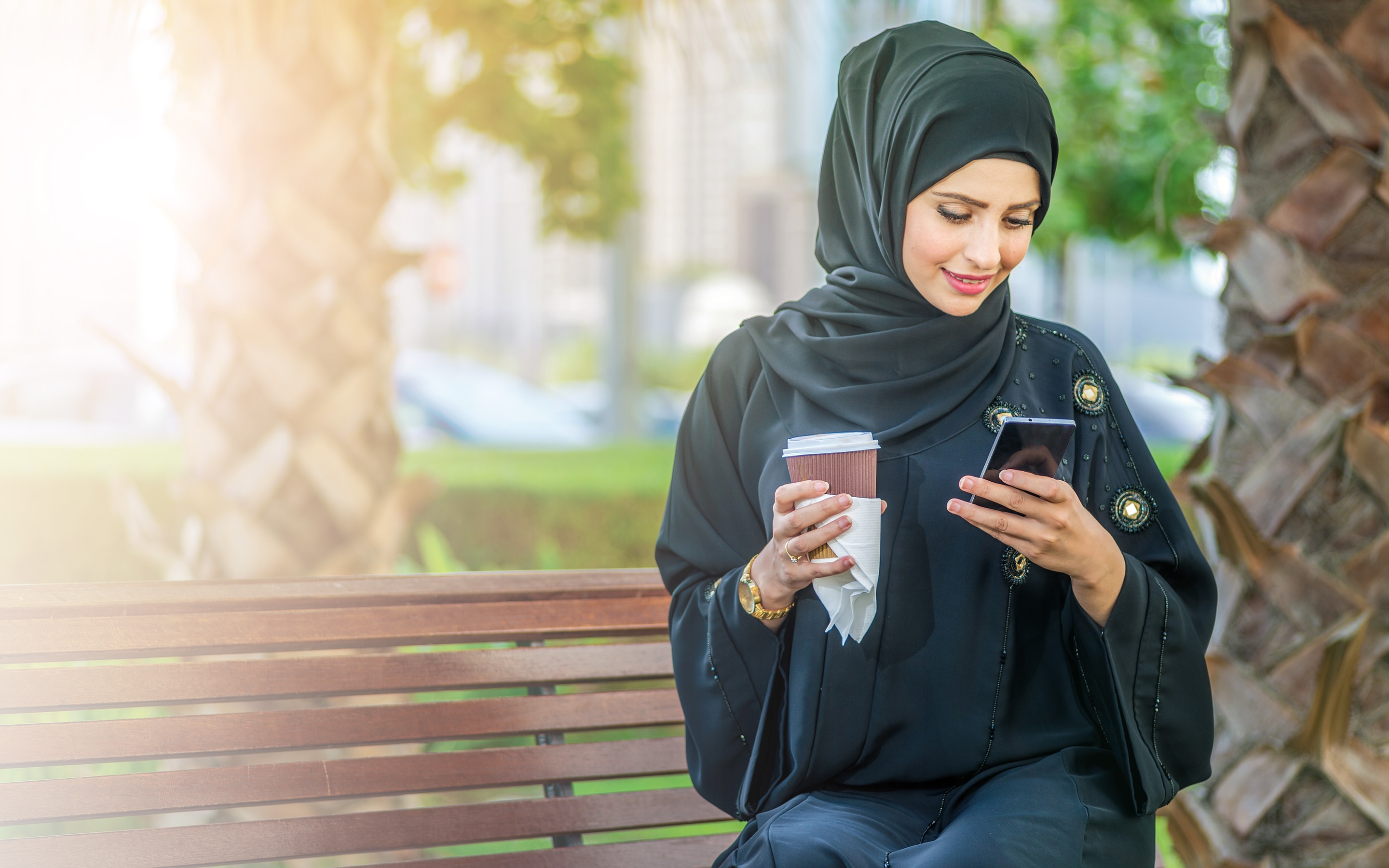  Arab businesswomen in hijab holding coffee outdoors and reading a message on a cell phone and looking at the camera.