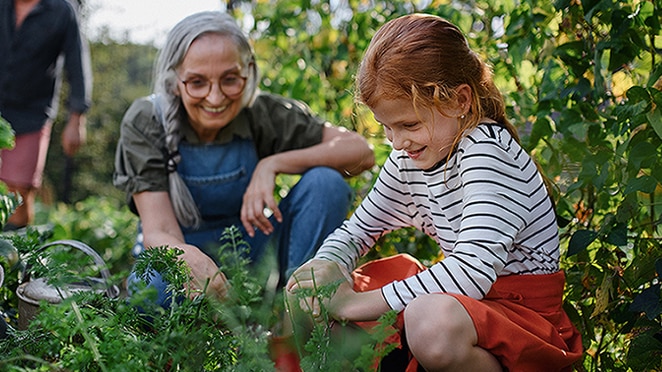 Older woman in overalls kneeling while watching a young child in the garden and smiling