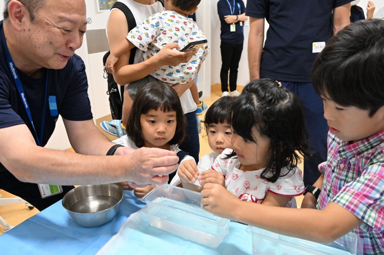 メドトロニックの従業員と共に、人工の心臓弁実物を触って体験中の子供たち