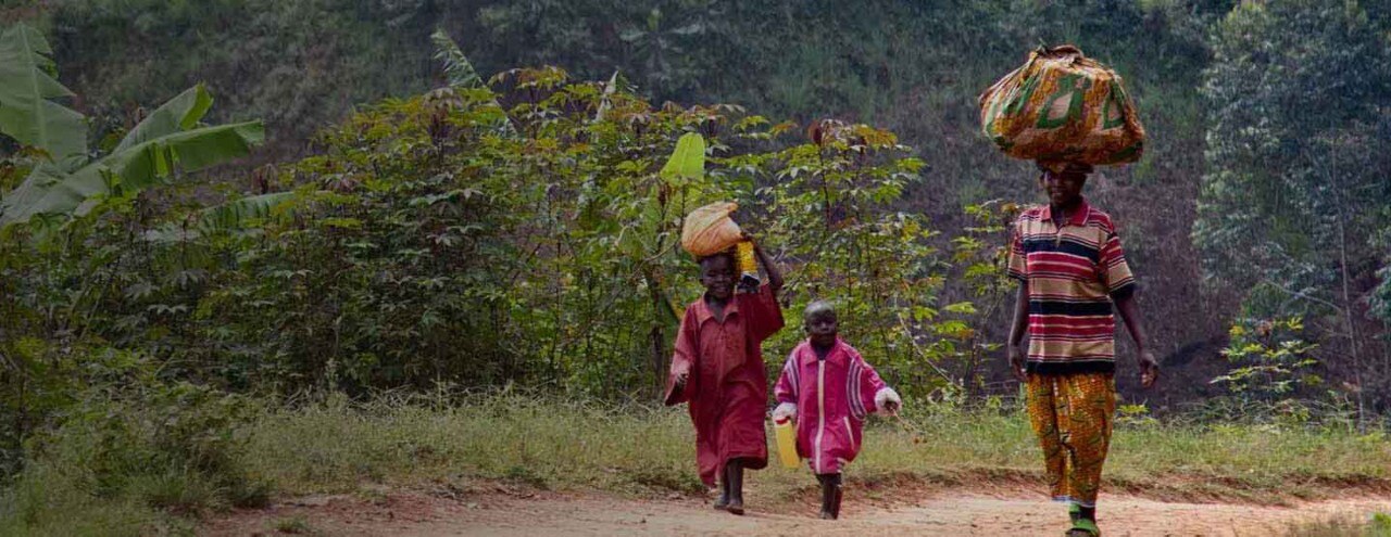 Three people carrying supplies walking on dirt road in green forest