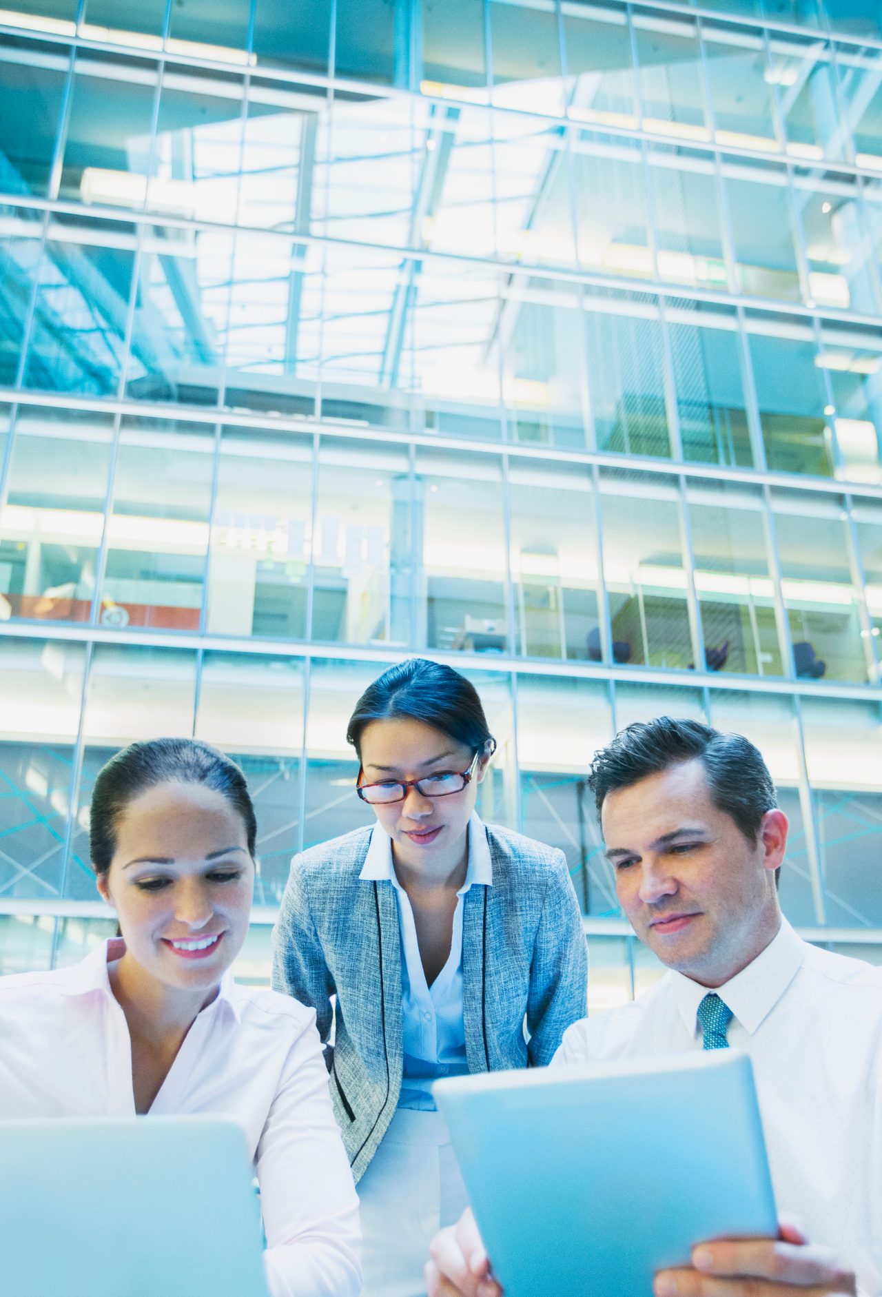 Two females and one male business professionals looking at a paper and a tablet in front of a glass building.